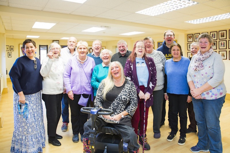 A group of people stood around a woman sat in a mobility scooter. Everyone smiling at the camera